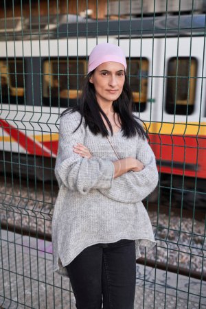 Téléchargez les photos : Jeune femme ethnique calme avec de longs cheveux foncés dans un pull chaud et un foulard rose debout près de la clôture avec les bras croisés et regardant la caméra sur la gare - en image libre de droit