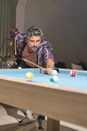 Photo for Serious middle aged bearded man with stylish haircut playing billiards in living room with cocktails and sofa on background - Royalty Free Image