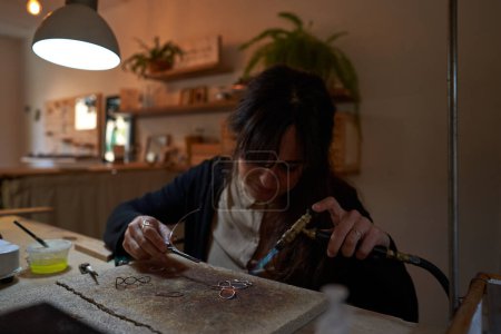 Foto de Artesana precisa concentrada con quemador de gas sentada en el banco de trabajo y haciendo accesorios hechos a mano en el taller - Imagen libre de derechos
