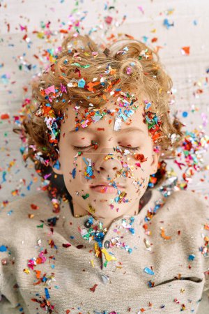 Téléchargez les photos : D'en haut d'adorable garçon préadolescent couché sur une surface blanche avec les yeux fermés et recouvert de confettis de poppers - en image libre de droit