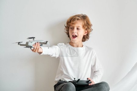 Foto de Niño contento con el pelo claro en ropa casual jugando con el dron de juguete mientras está sentado cerca de la pared blanca - Imagen libre de derechos