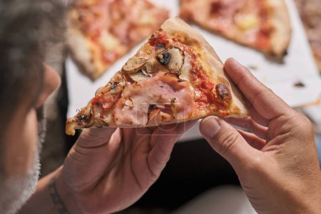 Téléchargez les photos : D'en haut de la culture personne anonyme manger morceau de délicieuse pizza maison à la maison - en image libre de droit