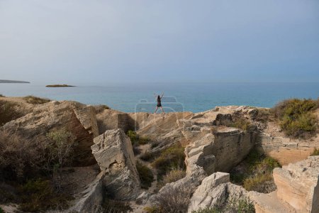 Téléchargez les photos : Vue de dos pleine longueur de coureuse anonyme debout sur le bord pierreux près de la mer sans fin calme et levant les bras tout en profitant de paysages étonnants - en image libre de droit