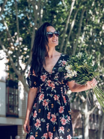 Téléchargez les photos : Charmante femelle aux longs cheveux foncés portant une robe d'été noire et des lunettes de soleil tenant un bouquet de fleurs en fleurs tout en marchant près des arbres dans la rue ensoleillée de la ville et en regardant ailleurs - en image libre de droit