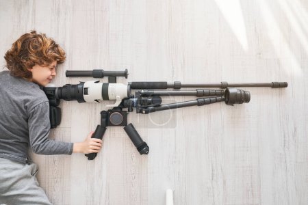 Photo for Top view of boy looking through scope of sniper rifle composed with equipment for photography while lying on wooden floor in light room during weekend day - Royalty Free Image