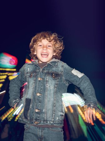 Téléchargez les photos : Garçon heureux en tenue décontractée regardant la caméra tout en sautant près de l'attraction éclairée dans le parc d'attractions public en soirée - en image libre de droit