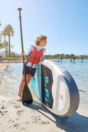 Téléchargez les photos : Corps entier d'un enfant positif portant un gilet de sauvetage debout près de la planche à pagaie sur une plage de sable lavé par une mer ondulante le jour d'été lumineux - en image libre de droit