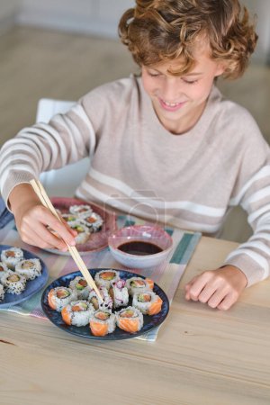 Téléchargez les photos : Angle élevé de garçon de culture souriant et regardant vers le bas tout en étant assis à la table en bois et en mangeant des rouleaux de sushi avec de la sauce soja en utilisant des baguettes en bois dans la cuisine à la maison - en image libre de droit