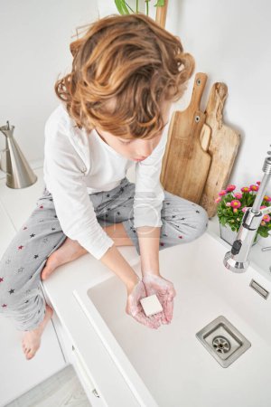 Photo for High angle full body of barefoot boy in pajama sitting on counter near cutting boards and washing hands with soap in kitchen in morning - Royalty Free Image