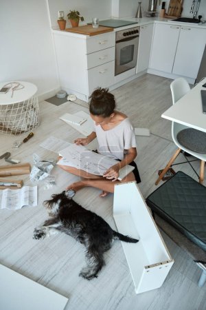 Photo for From above full body of barefoot woman sitting on floor with crossed legs near obedient purebred Miniature Schnauzer and reading furniture assembly instruction in light kitchen - Royalty Free Image