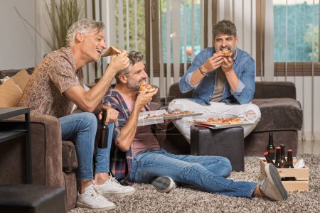 Foto de Grupo de amigos varones maduros comiendo sabrosa pizza y bebiendo cerveza mientras están sentados en la sala de estar en casa - Imagen libre de derechos