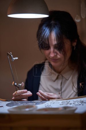 Foto de Maestro femenino concentrado trabajando con sierra en mesa iluminada por lámpara en estudio de joyería - Imagen libre de derechos
