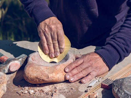 Téléchargez les photos : De haut de la récolte anonyme mâle artisan meulage de la roche avec du papier de verre tout en faisant de l'artisanat de pierre en plein air atelier - en image libre de droit