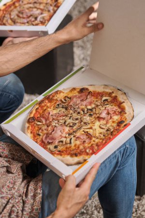 Téléchargez les photos : D'en haut des hommes de culture assis ensemble et ouvrir des paquets avec une pizza savoureuse tout en passant du temps libre à la maison - en image libre de droit