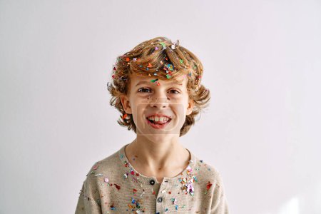 Photo for Cheerful cute preteen curly haired blond boy covered with colorful confetti and sparkles on white background - Royalty Free Image