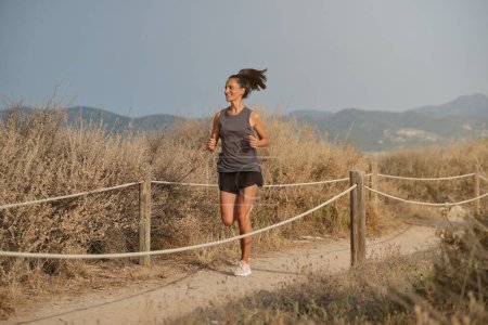 Téléchargez les photos : Corps entier d'athlète féminine souriante en vêtements de sport admirant les champs secs tout en faisant du jogging sur un sentier sablonneux contre les montagnes brumeuses - en image libre de droit