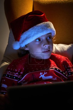 Photo for Charming boy in warm sweater and Santa hat with tablet relaxing in soft armchair while looking away in dark room with lamplight - Royalty Free Image