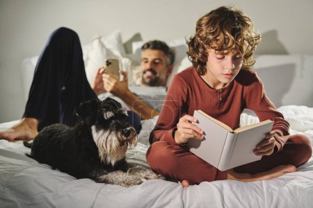 Téléchargez les photos : Père avec téléphone couché sur un lit confortable près du fils lisant un conte de fées intéressant près du chien miniature Schnauzer dans la chambre à coucher à la maison - en image libre de droit