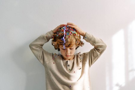 Téléchargez les photos : Triste garçon d "âge préscolaire aux cheveux bouclés avec tête tombante recouverte d" étincelles et de confettis sur fond blanc - en image libre de droit
