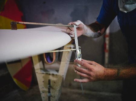 Photo for Crop anonymous male master in respirator measuring thickness of surfboard with ruler while shaping board placed in racks in workshop - Royalty Free Image