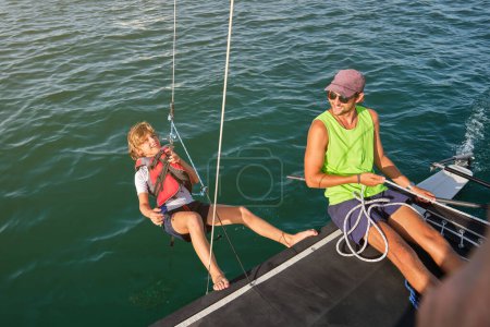 Photo for From above of cheerful young ethnic man in casual clothes and sunglasses smiling while pulling rope with fastened happy preteen boy during trip in sea on catamaran - Royalty Free Image