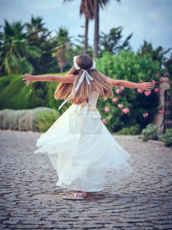 Photo for Back view of unrecognizable carefree preteen girl in white dress spinning around on path near green trees in summer garden - Royalty Free Image