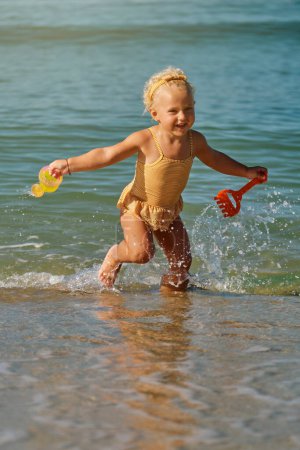 Téléchargez les photos : Adorable fille blonde jouant dans l'eau de mer sur la plage de sable avec arrosoir et pelle dans les mains seule pendant la journée - en image libre de droit