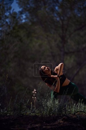Téléchargez les photos : Femme hispanique en vêtements de sport pratiquant la posture Parivritta Utkatasana avec les mains croisées pendant l'entraînement de yoga dans la nature le jour ensoleillé d'été - en image libre de droit
