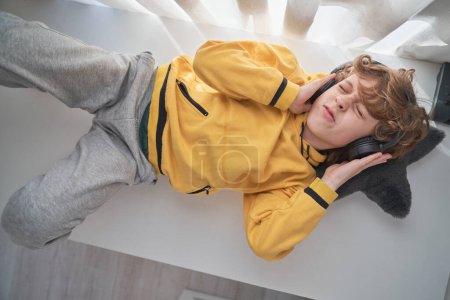 Téléchargez les photos : Vue aérienne du garçon de contenu en sweat à capuche jaune et casque couché sur la table à la maison et écoutant de la musique préférée - en image libre de droit