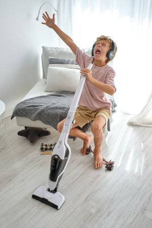 Photo for High angle of barefoot boy raising arm and yelling into vacuum cleaner while sitting on comfortable seat with cushions and blanket and cleaning floor in daytime at home - Royalty Free Image
