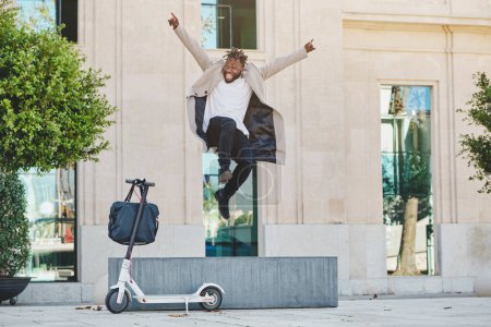 Téléchargez les photos : Ethnique heureux jeune homme en manteau sautant devant scooter électrique sur la chaussée en béton contre le bâtiment moderne en plein jour - en image libre de droit