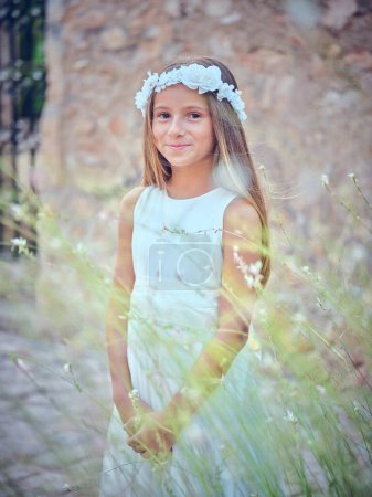 Photo for Calm young girl in white dress and floral wreath looking at camera while standing in grassy meadow in countryside - Royalty Free Image
