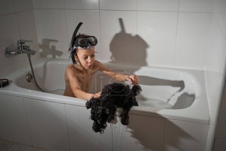 Téléchargez les photos : Garçon mignon avec torse nu dans le masque de plongée assis dans la baignoire avec chien noir pendant la routine quotidienne dans la salle de bain légère - en image libre de droit
