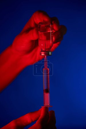 Foto de Persona irreconocible que demuestra jeringa y vial llenos de la vacuna COVID 19 y que se prepara para la inyección en un lugar oscuro sobre fondo azul - Imagen libre de derechos