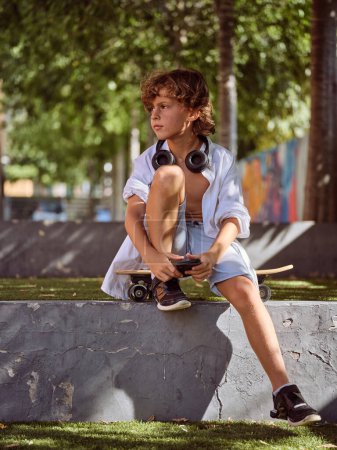 Foto de Cuerpo completo de niño preadolescente sentado en el monopatín en el parque con auriculares mientras mira hacia otro lado contra el fondo verde en un día soleado - Imagen libre de derechos