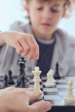 Téléchargez les photos : Crop personne méconnaissable jouer aux échecs à bord avec un garçon mignon sur fond flou tout en étant assis dans la pièce lumineuse à la maison - en image libre de droit