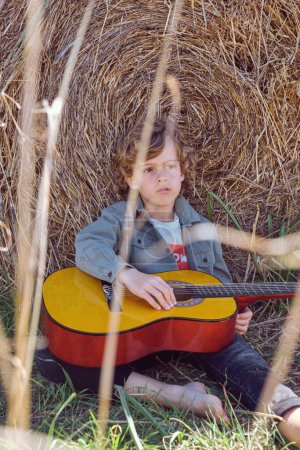 Téléchargez les photos : Garçon cher pieds nus avec les cheveux bouclés assis avec guitare sur l'herbe près de balle de foin dans le champ en zone rurale - en image libre de droit