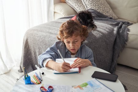 Téléchargez les photos : D'en haut du petit garçon écrivant dans un cahier assis près du canapé avec un chiot et faisant ses devoirs à la maison - en image libre de droit