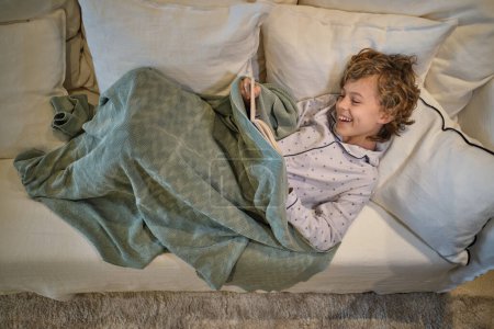Foto de Desde arriba cuerpo completo de niño alegre en pijama libro de lectura mientras descansa en cómodo sofá en la sala de estar de luz - Imagen libre de derechos