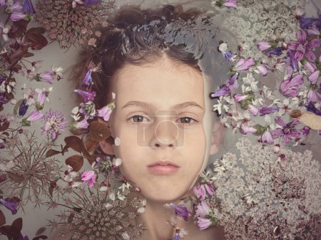 Téléchargez les photos : Vue de dessus de mignon garçon touchant les cheveux et regardant la caméra tout en étant couché dans une baignoire propre avec des fleurs colorées dans la salle de bain - en image libre de droit