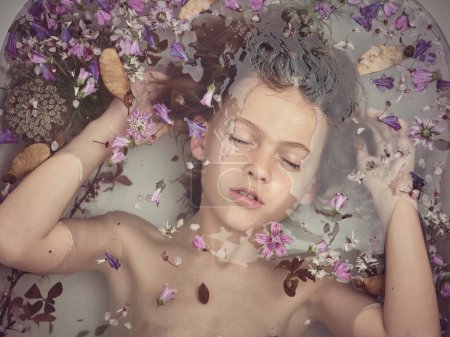 Photo for Top view of cute boy with closed eyes touching hair while lying in clear water of tub with colorful flowers in light bathroom - Royalty Free Image
