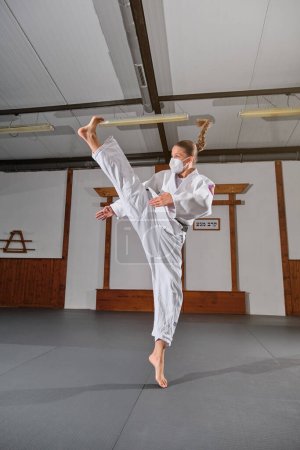 Photo for Full body of young female in kimono and medical mask practicing Krav Maga in gym room - Royalty Free Image