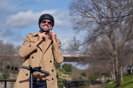 Foto de Hombre activo positivo en abrigo que se pone el casco protector mientras está de pie en scooter en el parque con árboles sin hojas en el soleado día de primavera - Imagen libre de derechos