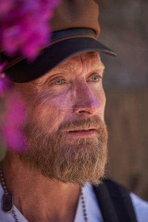Téléchargez les photos : Homme d'âge moyen réfléchi avec les yeux bleus et la moustache tout en se tenant près de fleurs violettes sur fond flou et en regardant loin le jour d'été dans la nature - en image libre de droit