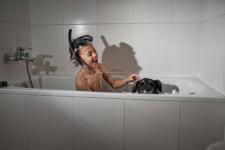 Foto de Vista lateral de niño alegre en máscara de buceo jugando con perro lindo en el baño durante la rutina diaria en baño ligero - Imagen libre de derechos