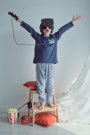 Foto de Longitud completa de niño alegre anónimo con la boca abierta levantando brazos mientras que regocija el éxito en el juego de realidad virtual con auriculares y el uso de controlador y auriculares contra el fondo blanco - Imagen libre de derechos