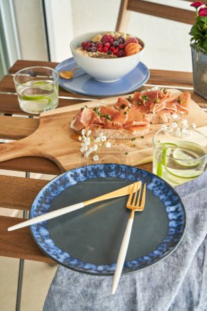 Téléchargez les photos : Assiette en céramique grand angle avec couverts servis sur la table près des toasts au prosciutto et bol de muesli et baies sur la terrasse - en image libre de droit