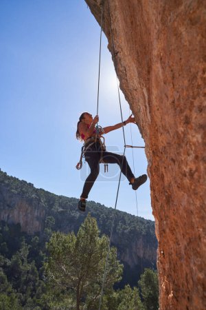 Foto de Bajo ángulo de la joven deportista con arnés de seguridad y ropa deportiva colgando del acantilado en la cuerda de escalada unida a la carabina en un día soleado - Imagen libre de derechos