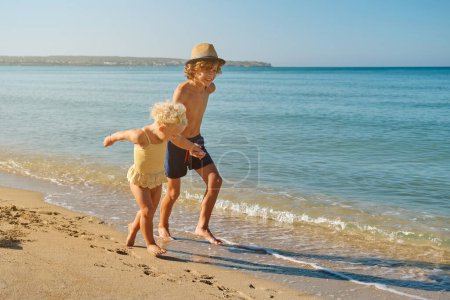 Téléchargez les photos : Délicieux préadolescent frère portant chapeau et maillots de bain tenant la main de petite sœur tout en courant ensemble sur la plage de sable près de la mer pendant les vacances d'été - en image libre de droit