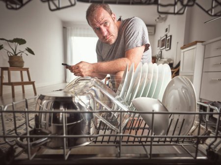 Téléchargez les photos : Message texte masculin sérieux et regardant la caméra tout en se tenant près du lave-vaisselle ouvert moderne avec vaisselle propre dans la cuisine légère - en image libre de droit